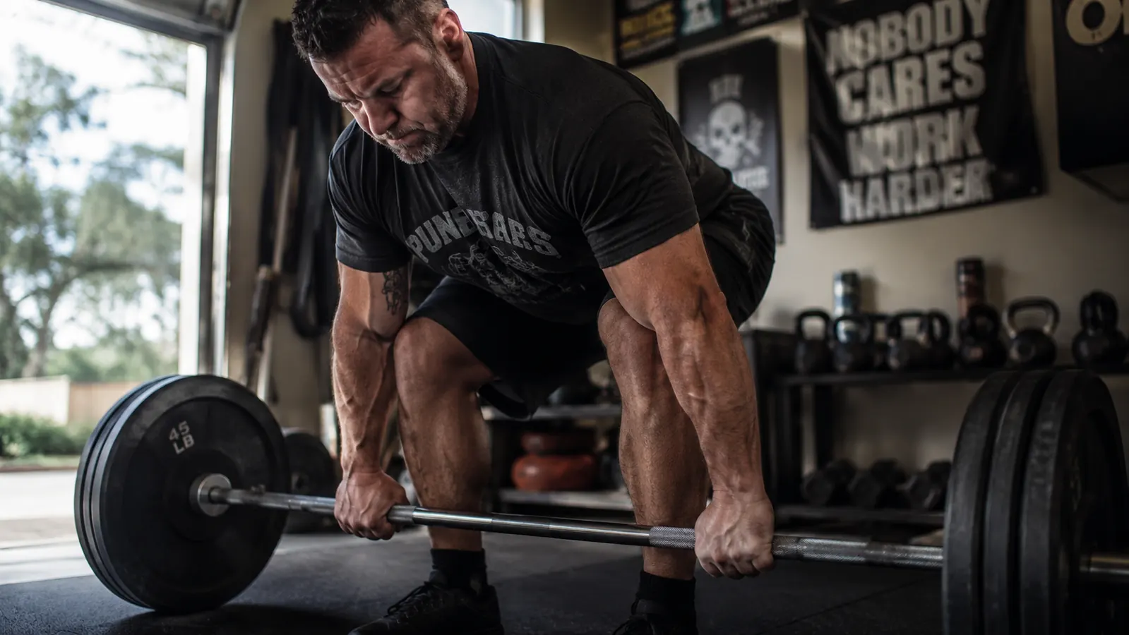 Man in his 40s finishing a set of deadlifts in a home gym, gripping the bar with visible forearm tendon definition