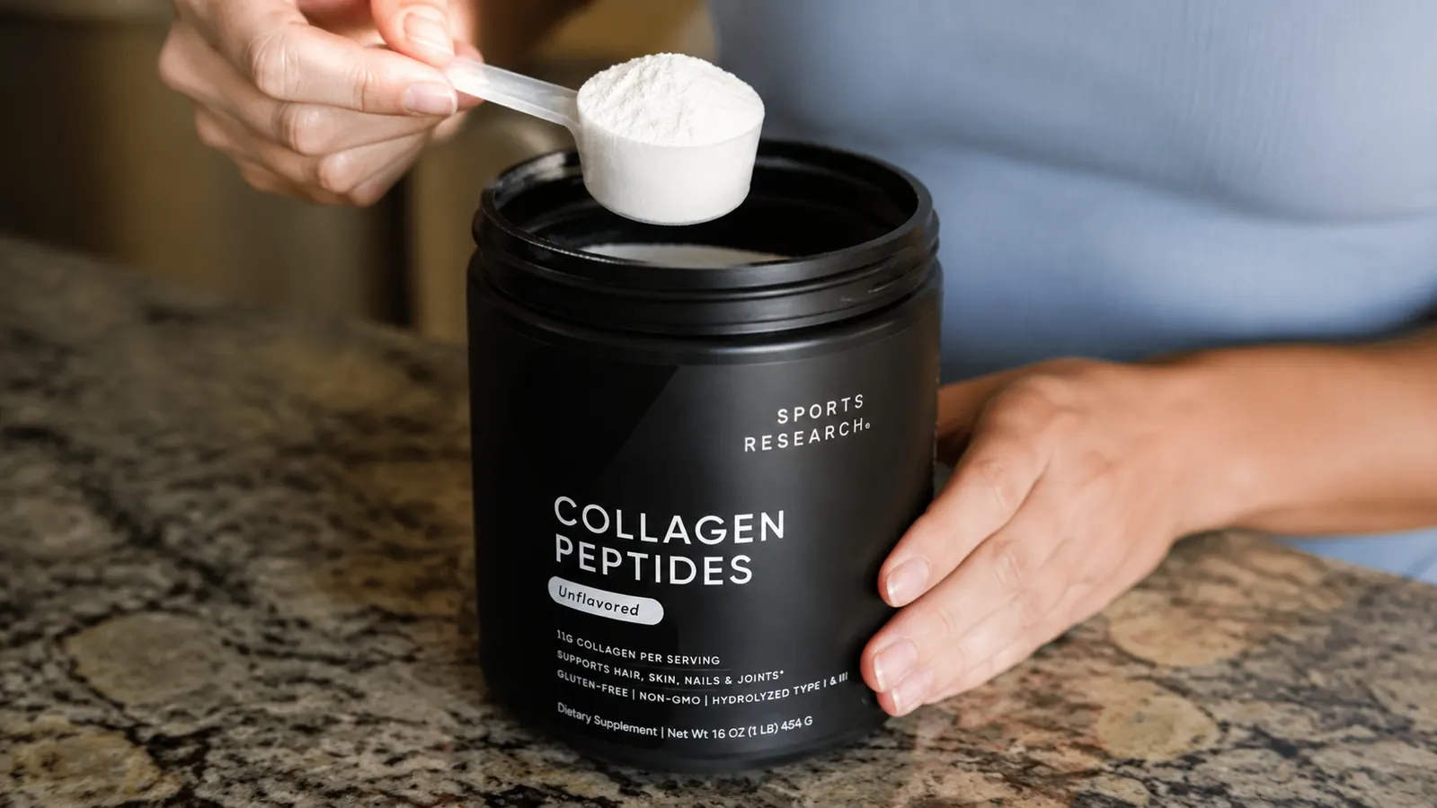 Hand scooping collagen peptides powder from the tub with a measuring scoop next to a glass of water